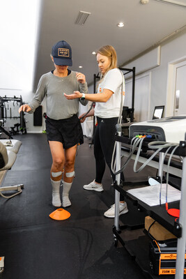 A physical therapist guides a patient through balance exercises using the Neubie device, with electrodes placed on the patient’s leg to enhance stability and proprioception. The therapist stands close by for support while the device delivers electrical stimulation to aid neuromuscular re-education.